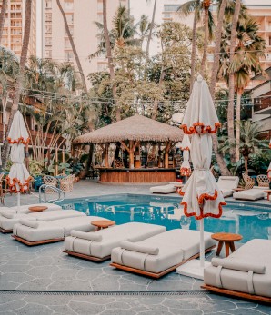 A tranquil poolside scene with lounge chairs, umbrellas, a tiki bar, and lush greenery in a resort setting.