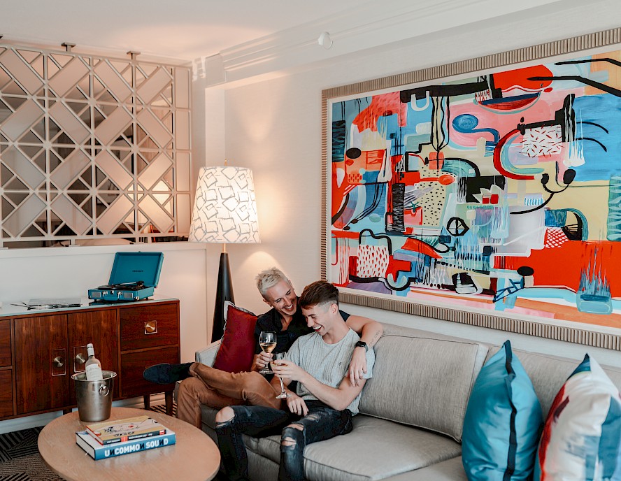 Two people sit on a couch in a modern living room with vibrant artwork, a turntable, and a lit lamp, sharing a drink and smiling.