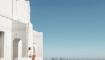A person sits on the edge of a white building, overlooking a sprawling cityscape under a clear blue sky. The city