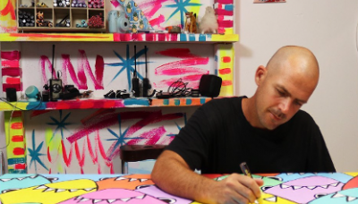 A person is painting a colorful, cartoonish artwork with monster faces. The background contains colorfully painted shelves and art supplies.