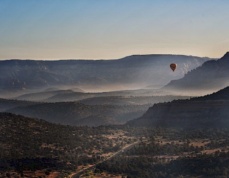 A hot air balloon floats over a scenic, mountainous landscape with mist settling in the valleys and the morning sun casting soft light.