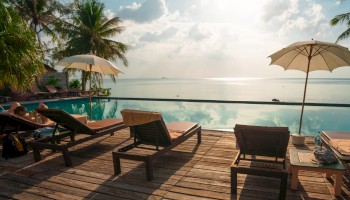 A serene poolside scene with lounge chairs, umbrellas, and a beautiful ocean view under a sunny sky, surrounded by lush palm trees.