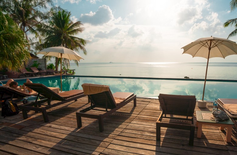 A tranquil poolside scene with lounge chairs, umbrellas, and a scenic view of the ocean and palm trees during a peaceful, sunny day.