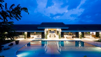 A luxurious modern building with an illuminated swimming pool at night, surrounded by seating and greenery, under a blue sky with some clouds.