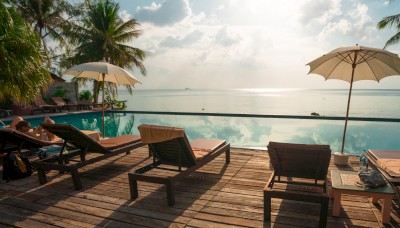 Lounge chairs, umbrellas, and palm trees by a pool overlooking a serene body of water under a partly cloudy sky, with the sun shining brightly in the distance.