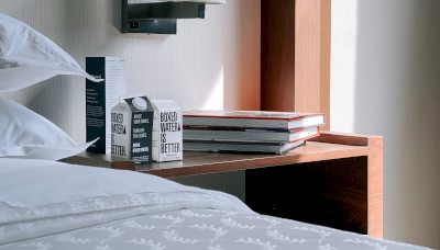 A bedside table with books, a carton of boxed water, and a light fixture above. The bed has pillows and a comforter with a leaf pattern.