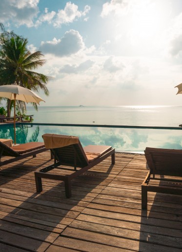The image shows a serene poolside with lounge chairs, umbrellas, palm trees, and a calm ocean view under a sunny, partially cloudy sky.