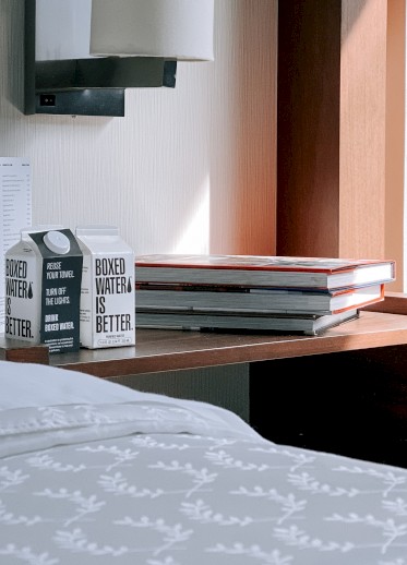 A bedside table with books, a carton of boxed water, and a modern wall lamp are next to a neatly made bed with white pillows and a patterned blanket.
