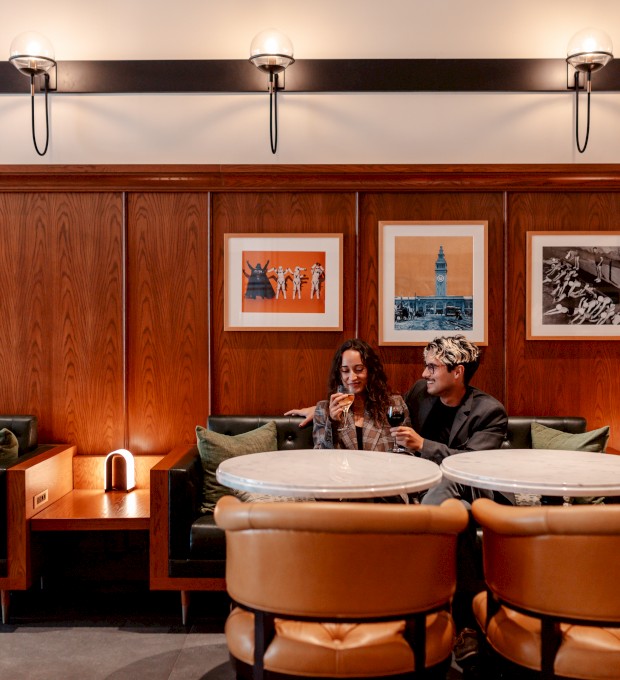 A cozy restaurant interior with two people sitting at a round table, enjoying drinks, surrounded by wood-paneled walls and framed pictures.
