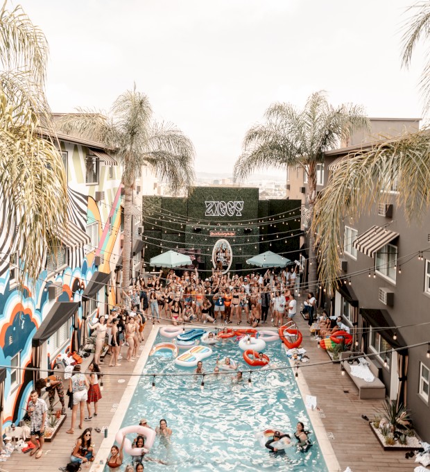 A lively pool party is taking place between two buildings decorated with murals; people are swimming and lounging around under umbrellas and palm trees.