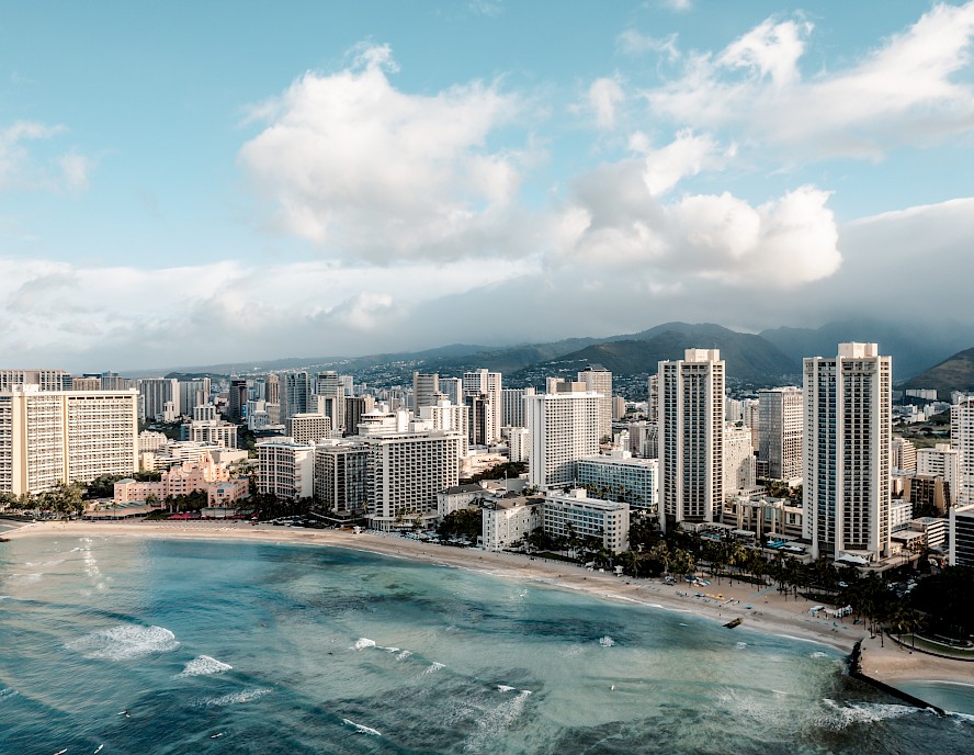 This image depicts a coastal cityscape with numerous high-rise buildings, a sandy beach, and a beautiful ocean with waves under a partly cloudy sky.