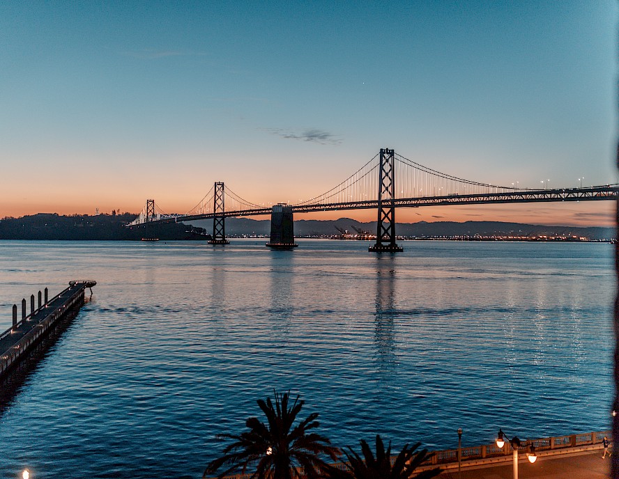 A scenic view of a bay with a large suspension bridge at sunset, including a pier, palm trees, and calm waters reflecting the sky