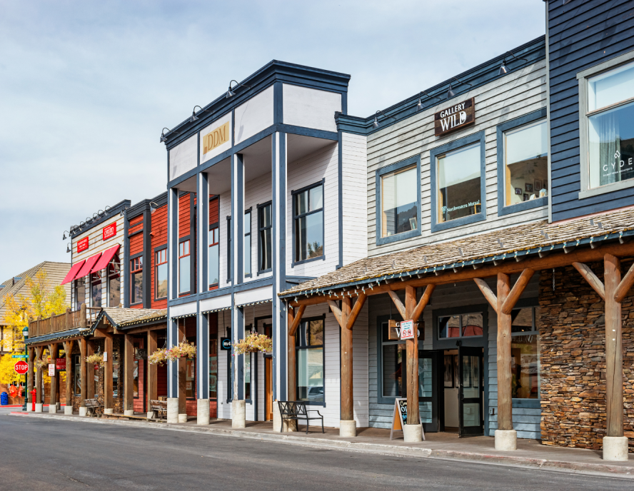 A street with western-style buildings, featuring a hotel on the left, shops, and wooden walkways. The scene is bright and inviting under a clear sky.