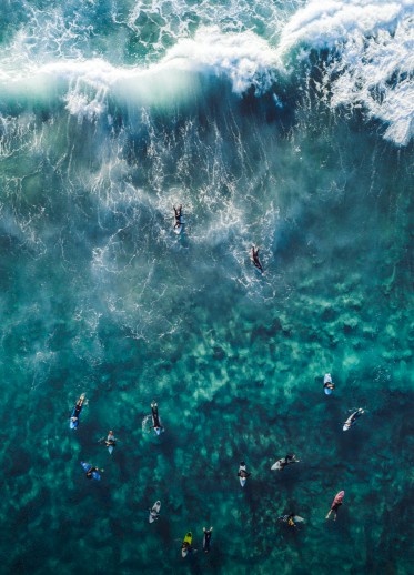 Aerial view of people surfing or swimming in a clear blue ocean, with larger waves at the top of the image, capturing the dynamic water motion.