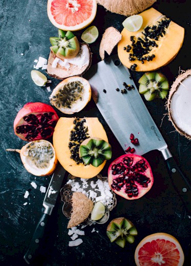 The image features assorted sliced fruits, including grapefruit, kiwi, passion fruit, and pomegranate, alongside two knives on a dark surface.