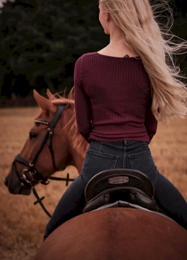 A person with long blonde hair, wearing a red sweater, is riding a brown horse through a field, viewed from behind.