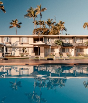 This image shows a serene pool area in front of a low-rise building surrounded by palm trees, with clear blue sky above and poolside umbrellas.