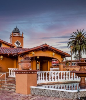 A picturesque building featuring Spanish-style architecture, with red-tiled roofs, outdoor staircases, potted plants, a fountain, and a palm tree.
