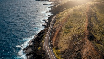 A winding coastal road with a few vehicles, running alongside a rocky shoreline with waves crashing, set against a backdrop of hills and a setting sun.