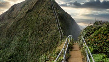 A narrow staircase winds up a green, steep mountainside under a dramatic, cloudy sky, offering a breathtaking and adventurous pathway.