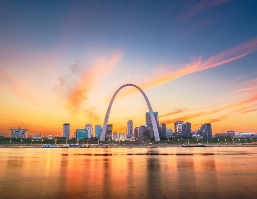 A scenic view of a city skyline at sunset, featuring a prominent arching structure near a waterfront, with colorful clouds in the sky.