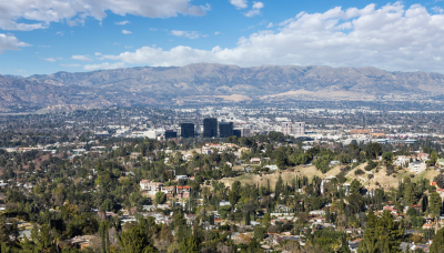A cityscape with residential areas, skyscrapers in the distance, and mountains under a partly cloudy sky, surrounded by greenery and trees.