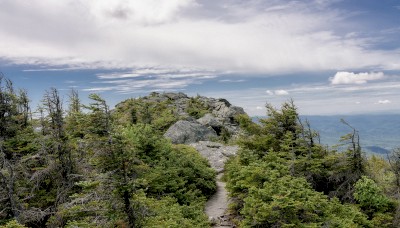 A rocky mountain trail surrounded by green trees leads to a scenic overlook with a partly cloudy sky in the background.